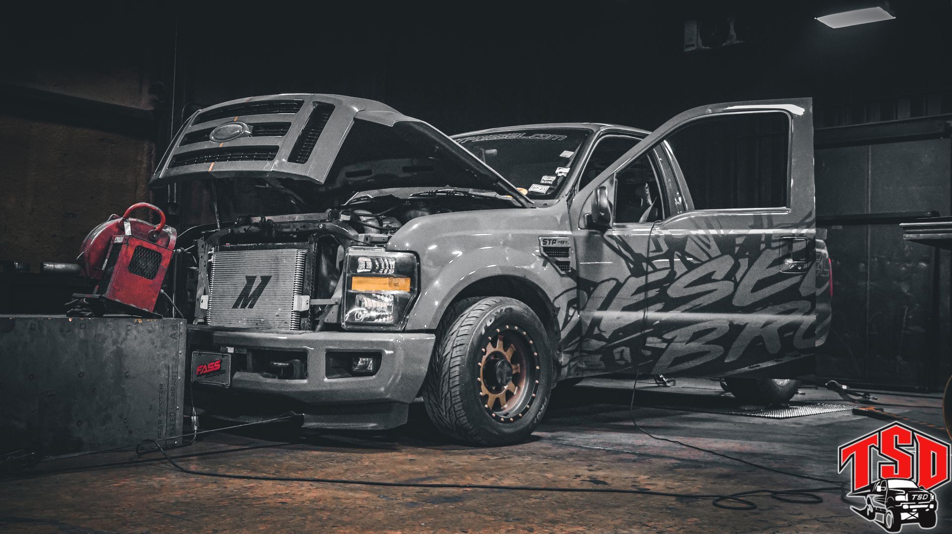 Modified silver pickup truck in a dim garage, hood and doors open, with red equipment nearby.
