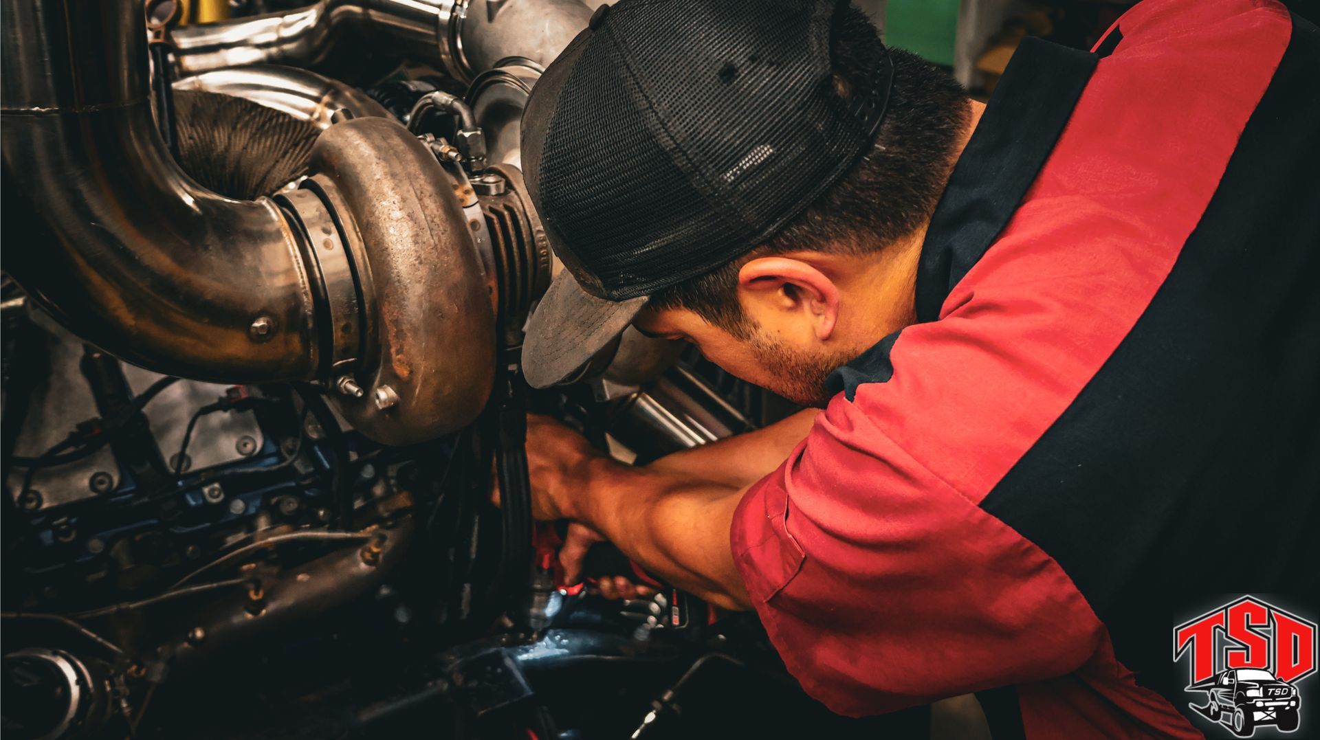 Mechanic in red shirt working under a vehicle, inspecting engine components in a dim garage.