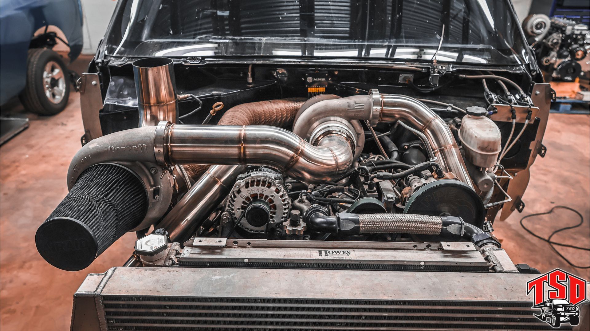 Classic car engine bay with polished custom exhaust tubing and large intake hose, front view