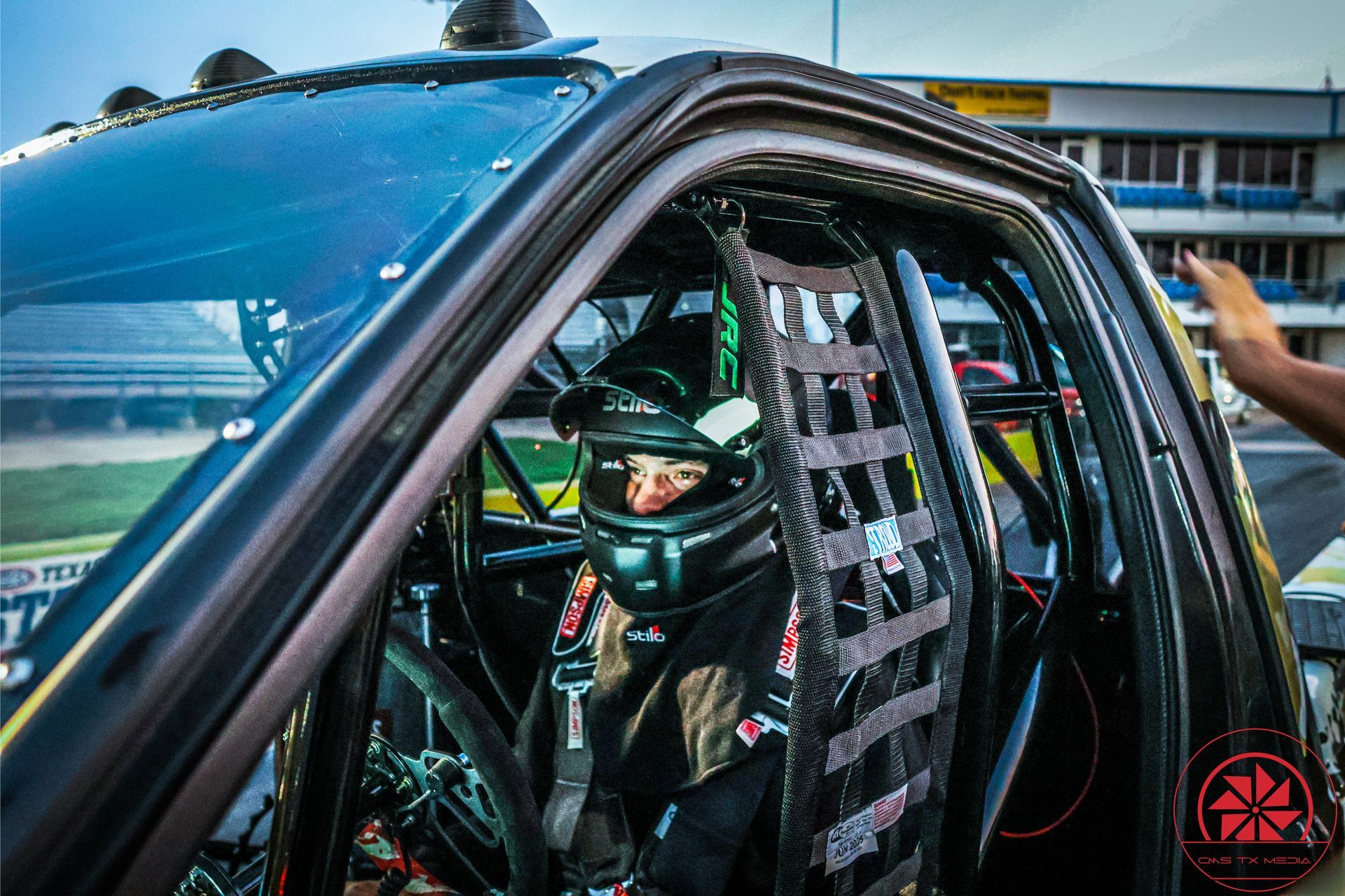 Race car driver in helmet and harness inside a blue car, seen through the open window.
