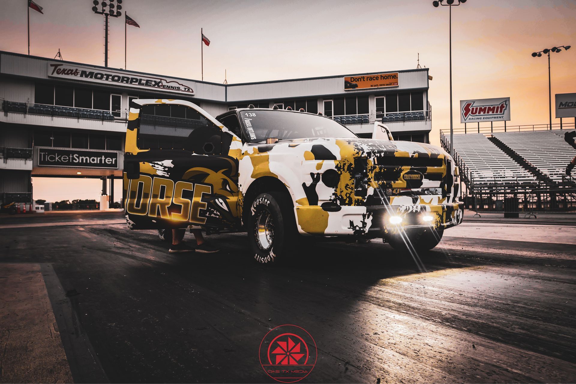 Camouflaged yellow-and-black car on a racetrack at dusk, headlights on, with stadium seating behind it