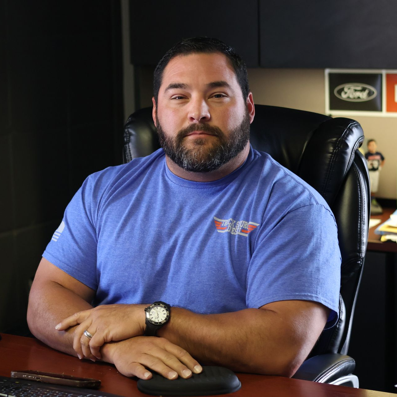 Chris Alaniz seated at a desk in an office, wearing a blue shirt and black watch, looking at the camera