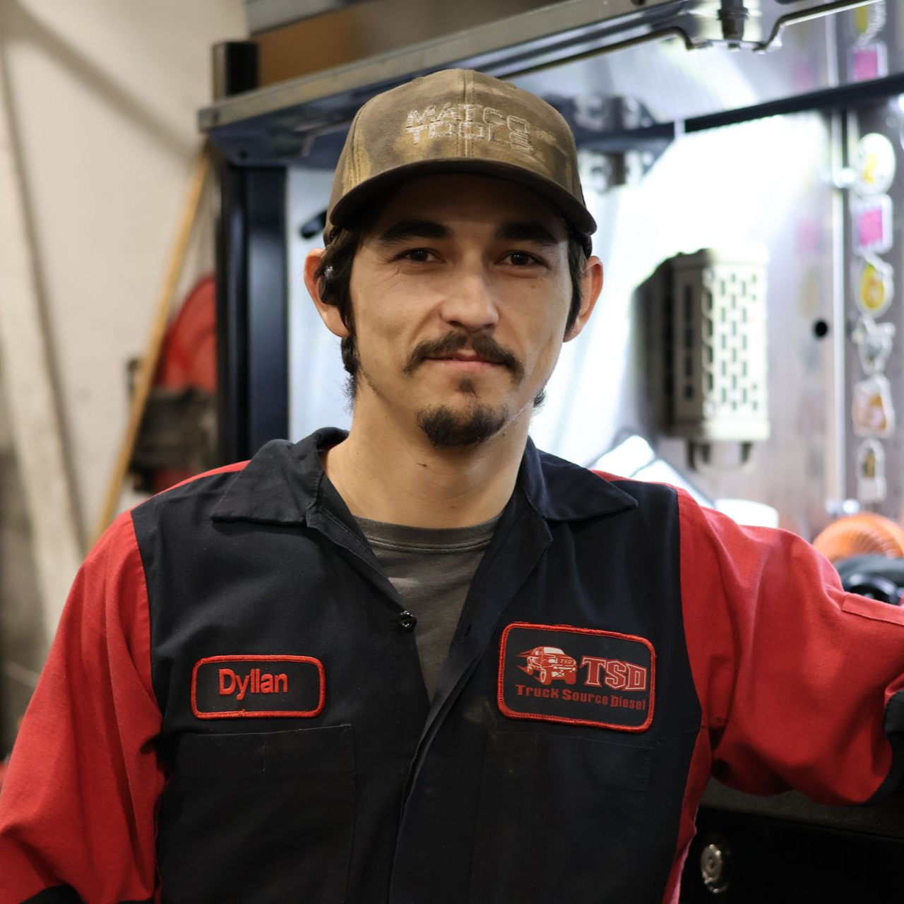 Dyllan Morin in red-and-black work vest and cap standing in a workshop, looking at the camera.