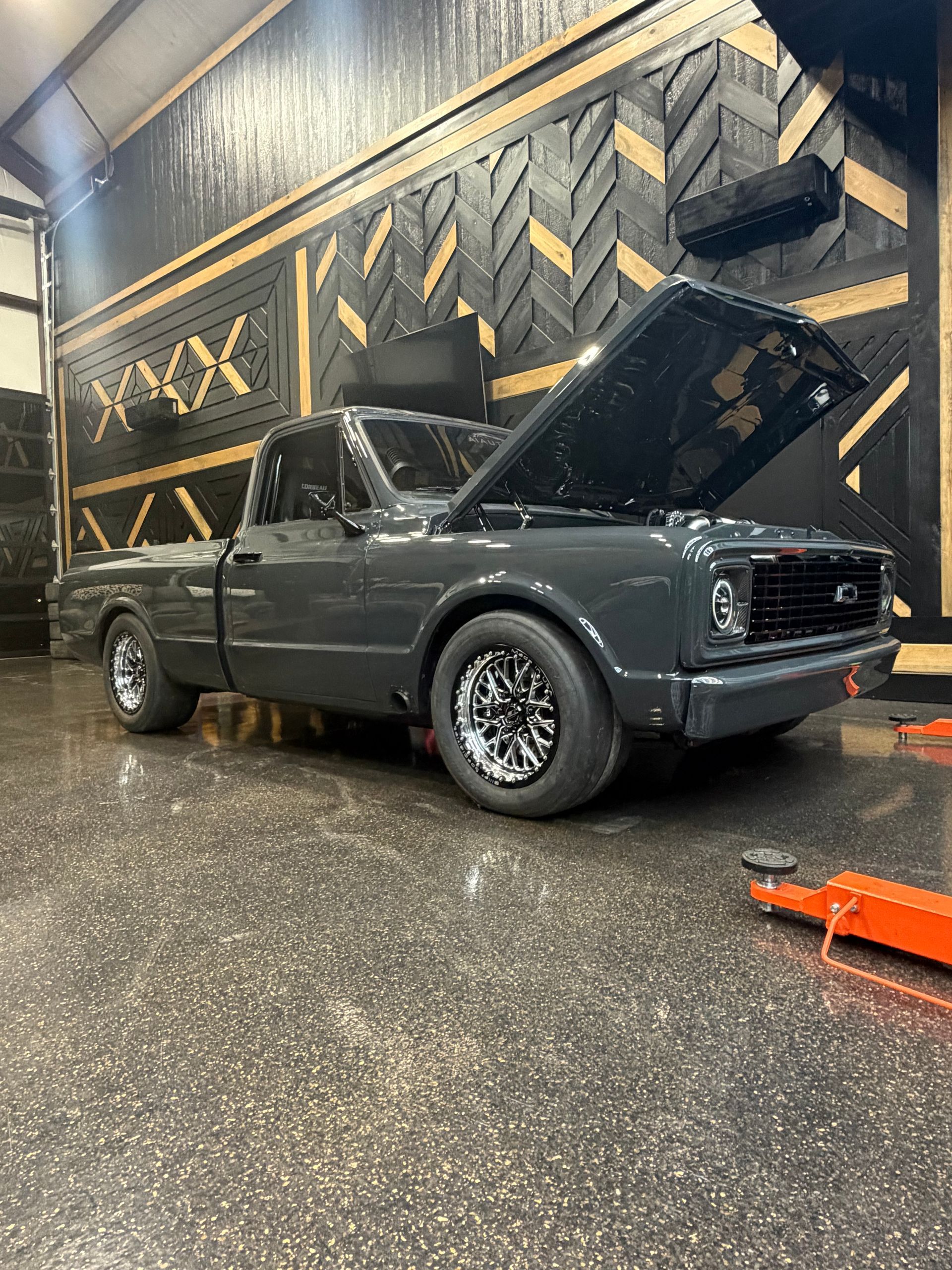 Classic dark gray pickup truck in a garage, hood open, with gold geometric wall art behind it.