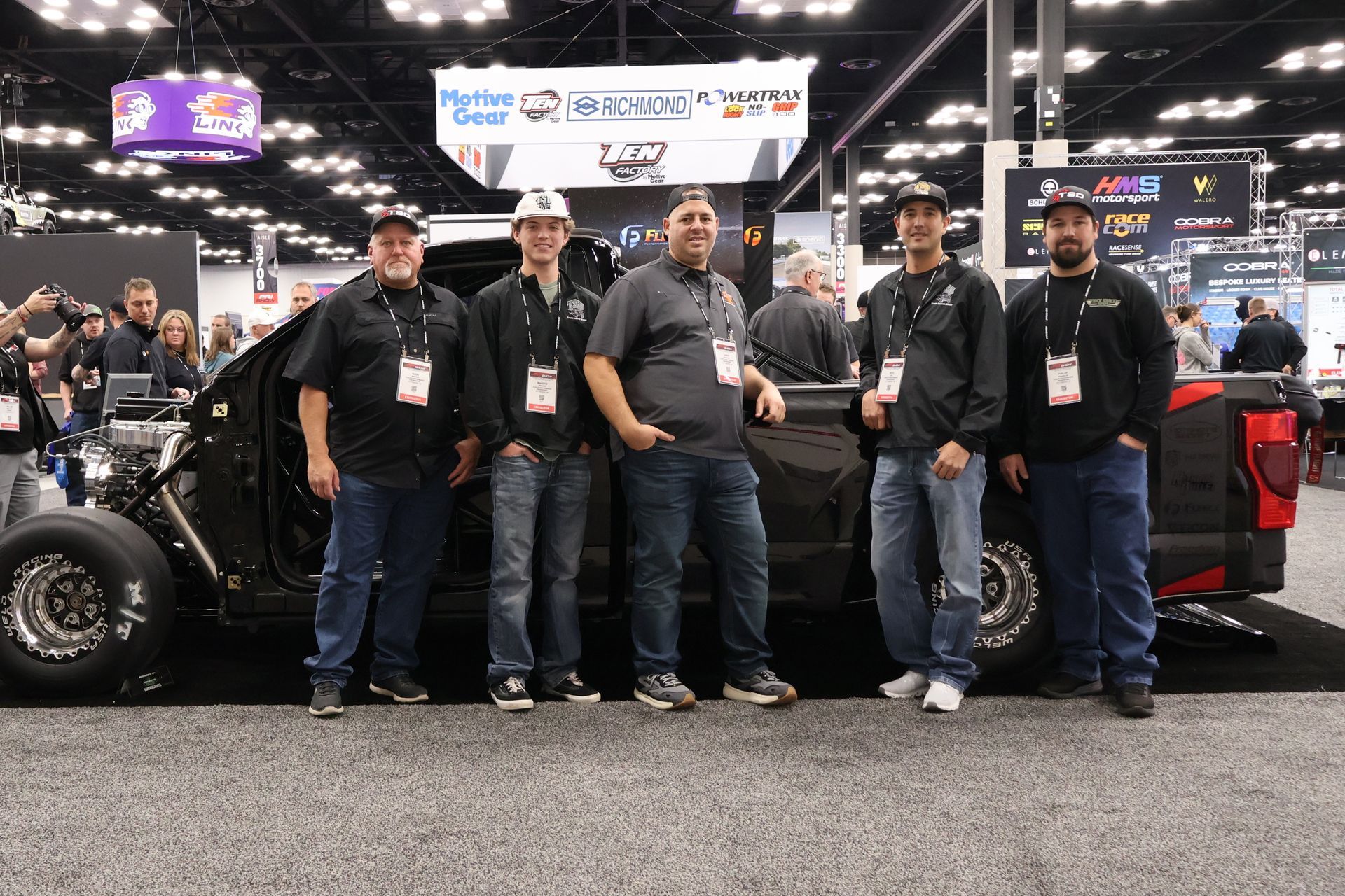 Five people standing in front of a black off-road vehicle at an indoor auto show display.