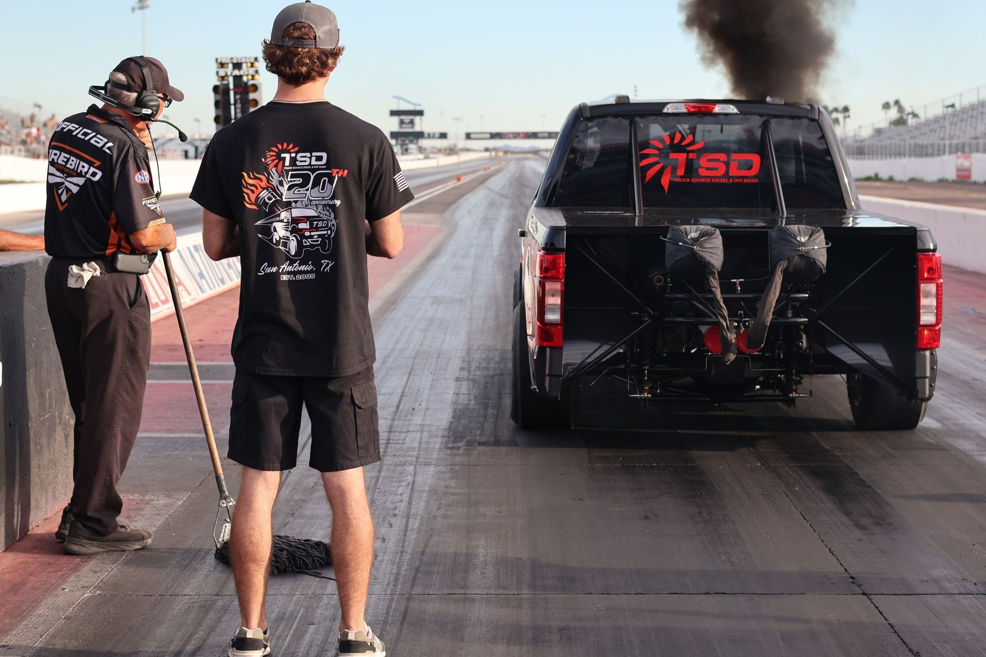 Two men near a black pickup truck on a drag strip, with smoke rising from the truck’s rear.