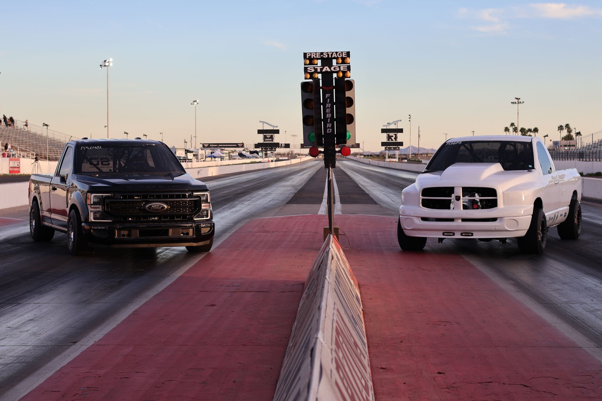 Two pickup trucks staged at a drag strip starting line at sunset, black truck left and white truck right.