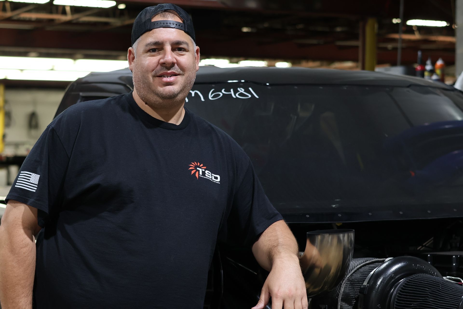 Chris Buhidar a black shirt and cap leaning on a Black diesel-powered race truck named Overnight
