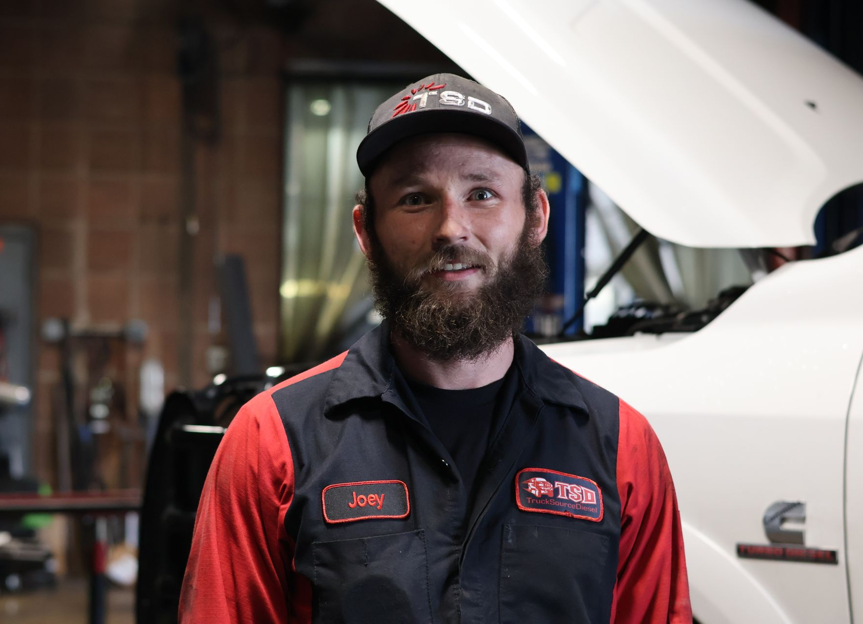 Joey Magnus standing in a garage beside a white truck with its hood open, wearing a black and red uniform.