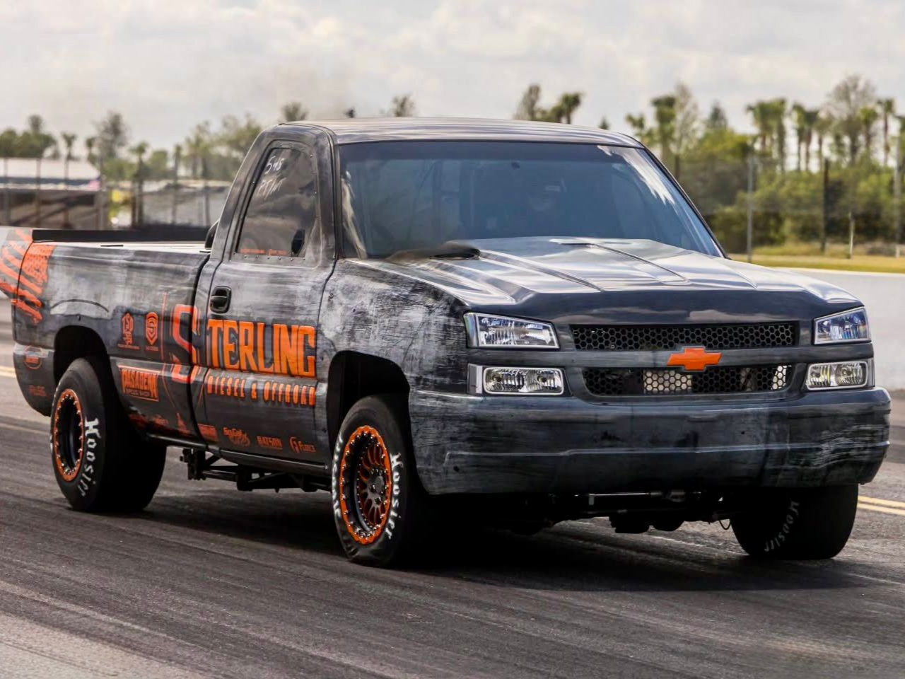 Black diesel-powered race truck named Overnight with orange decals speeding on a road, palm trees in background