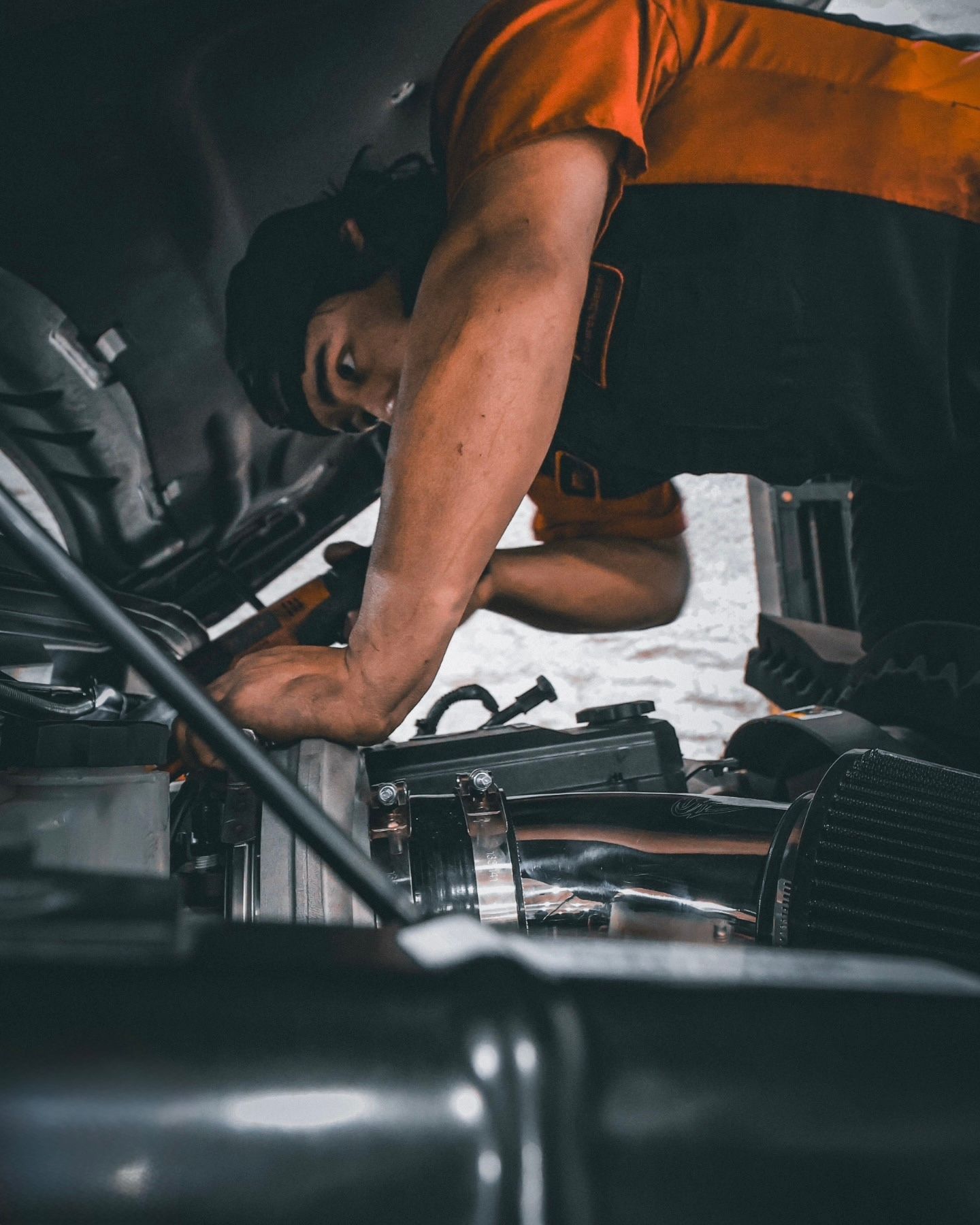 Mechanic in orange shirt working under a car hood in a garage