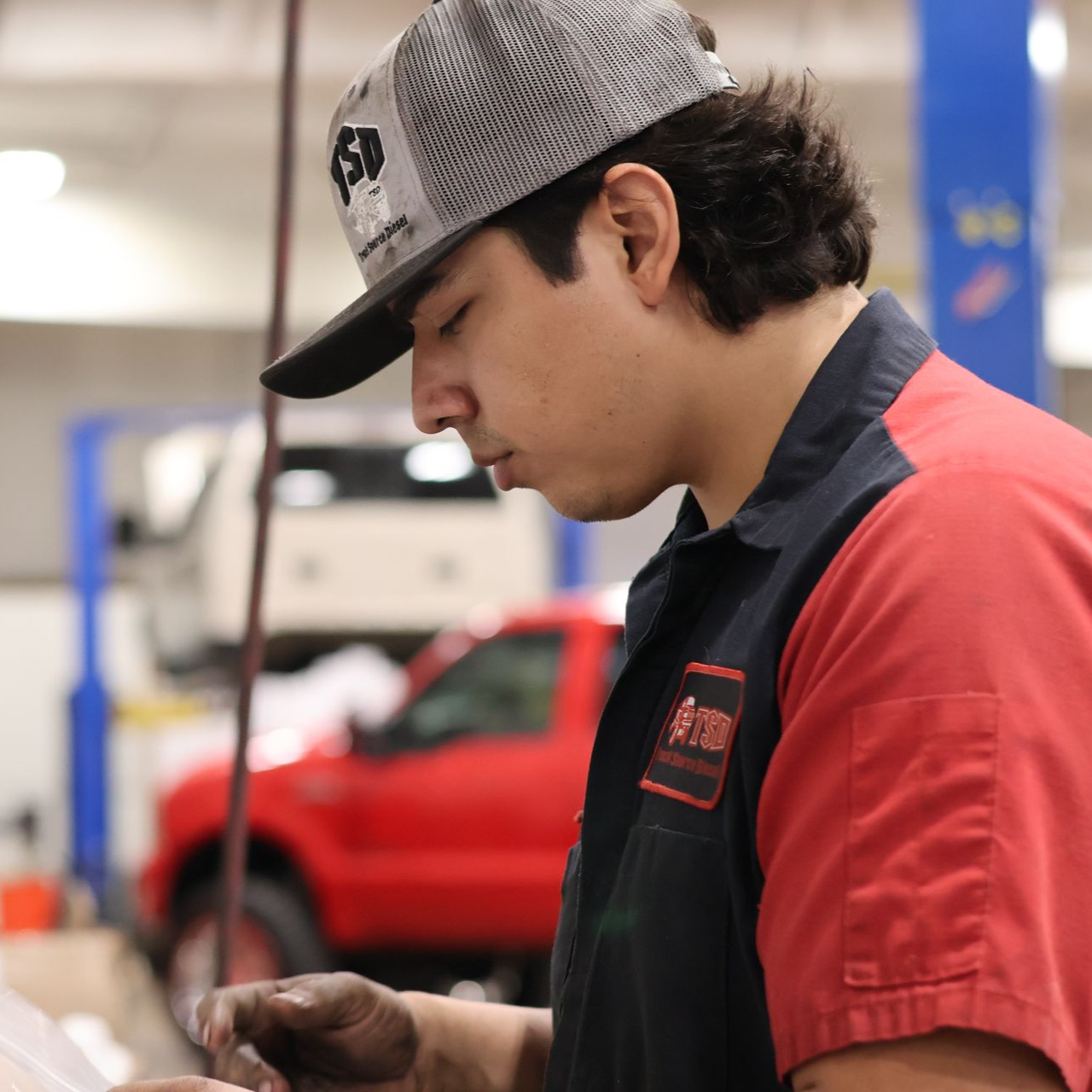 Manny Valenzuela in a gray cap and red shirt examining a small part in a garage, with a red car in the background