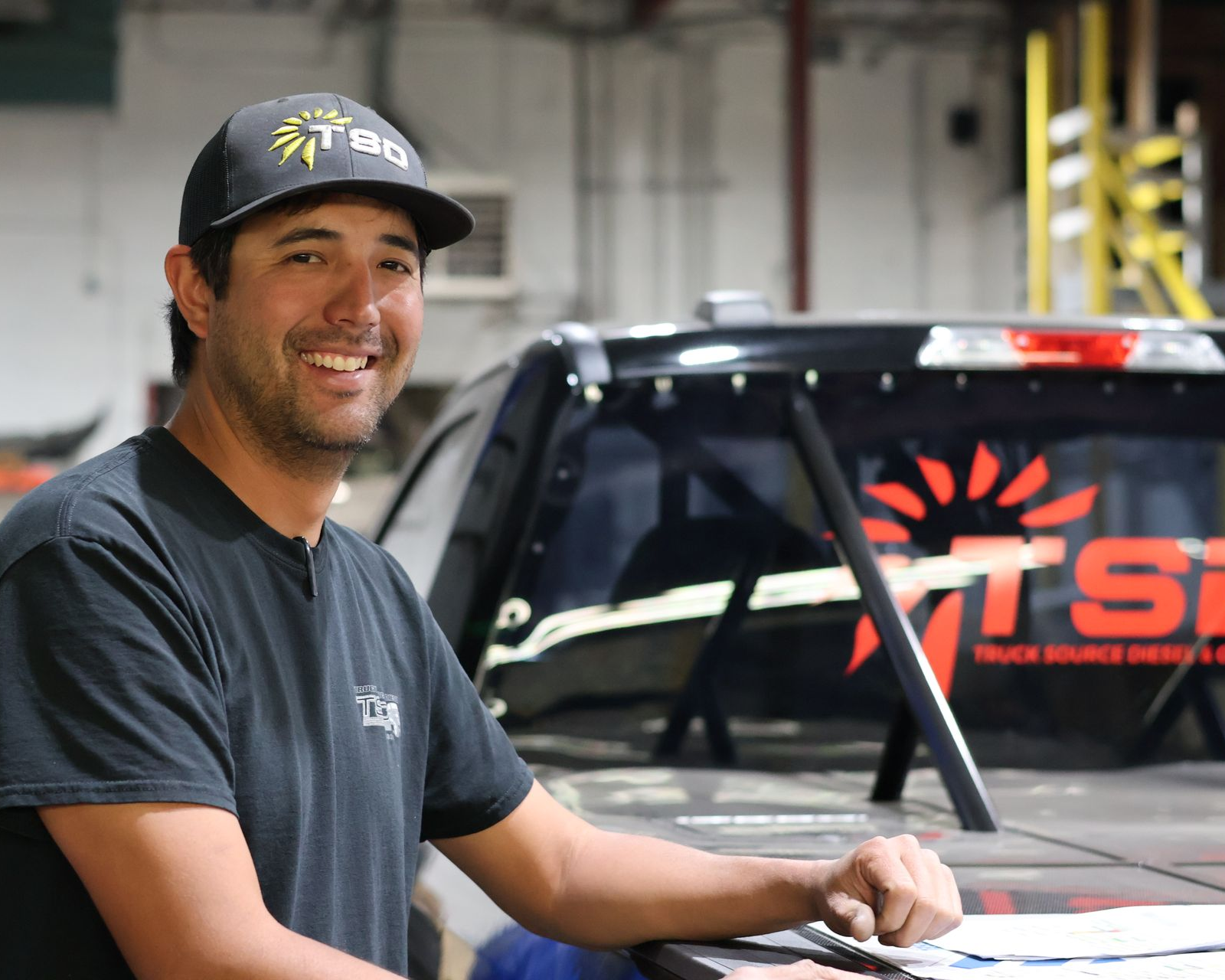 Nathaniel Oku in a black cap stands beside a Black diesel-powered race truck named Overnight