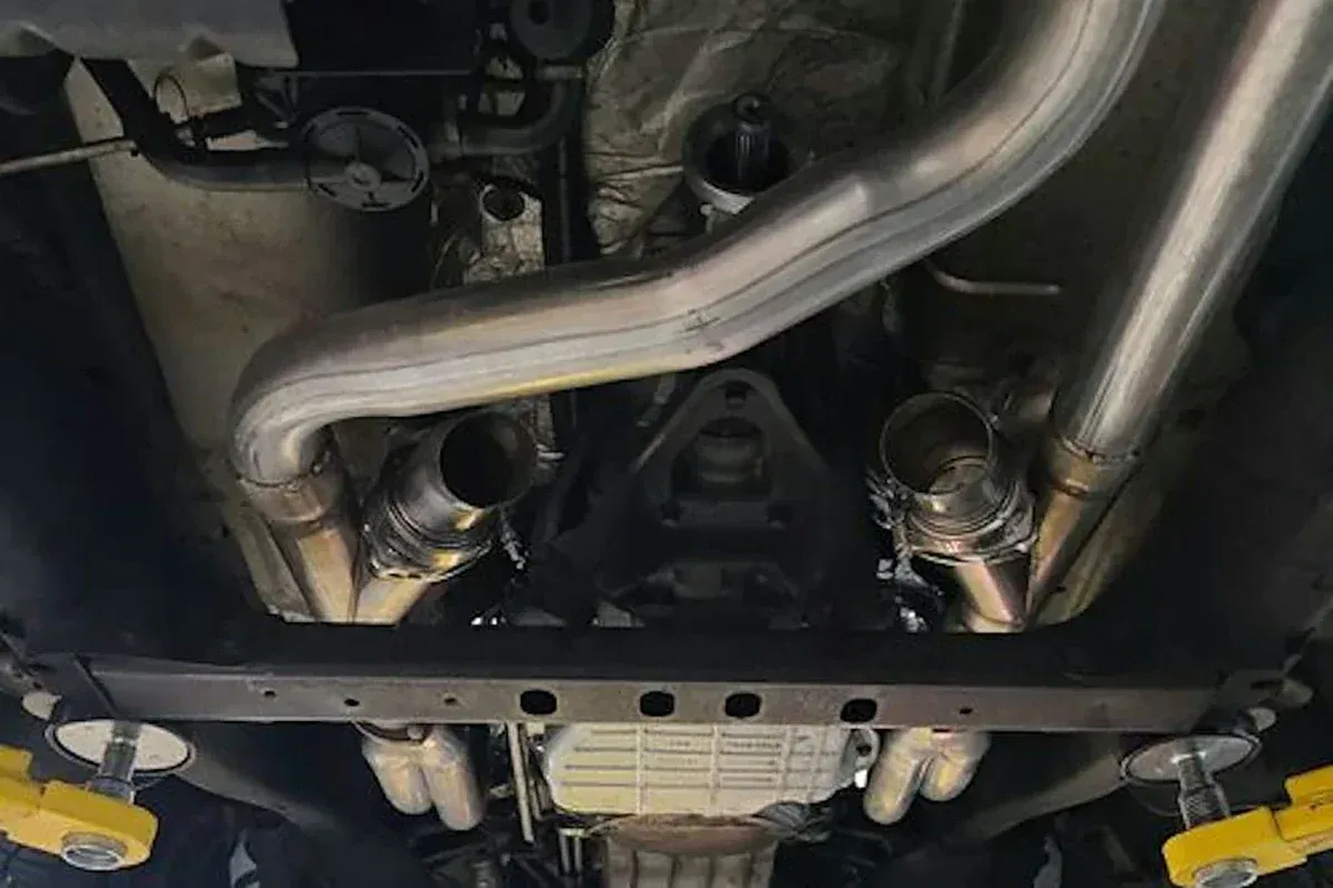 Undercarriage of a car showing shiny exhaust piping and a skid plate, with yellow lift arms visible below