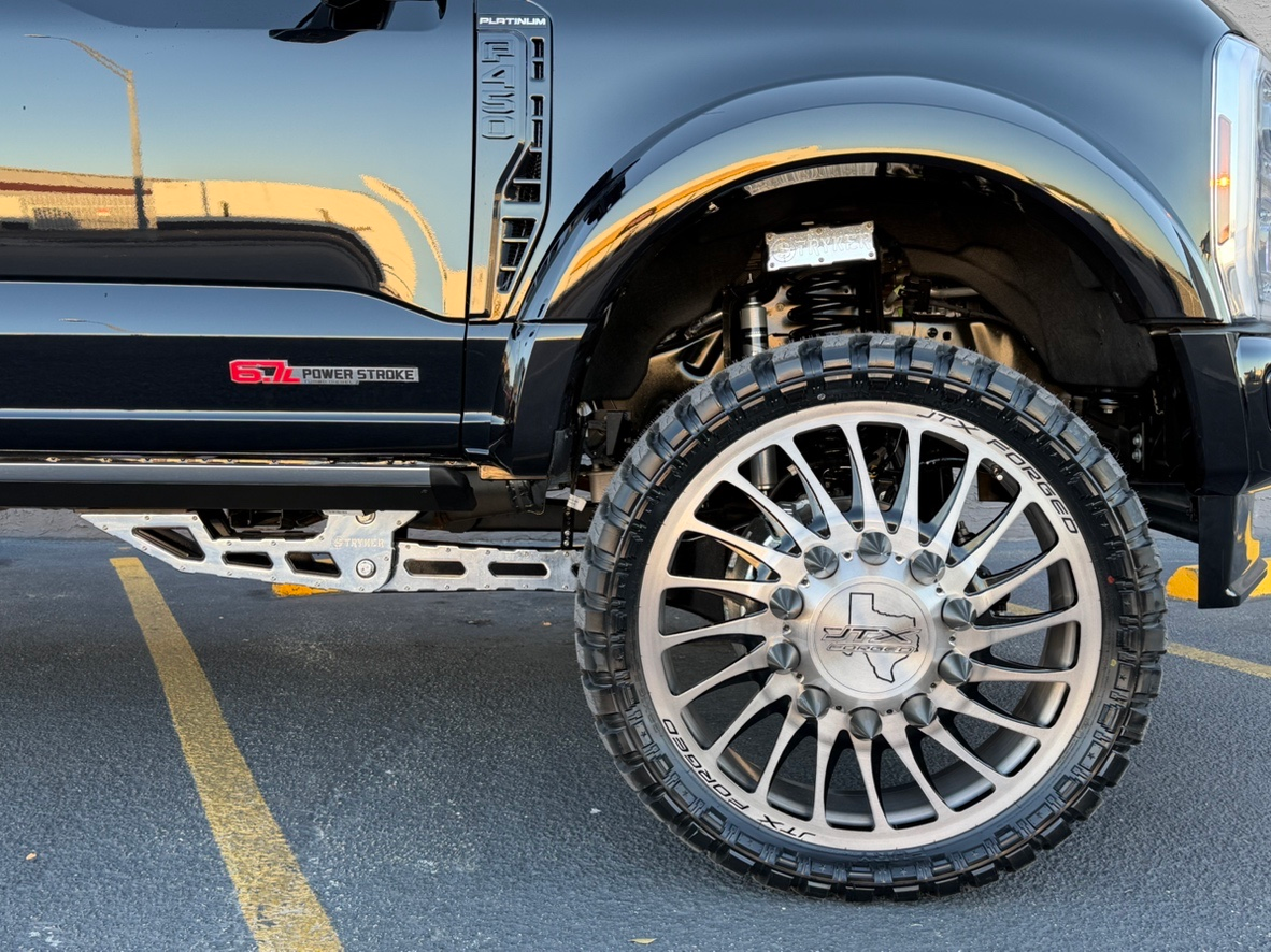 Black lifted pickup truck with oversized chrome wheel and off-road tire parked in a lot at sunset