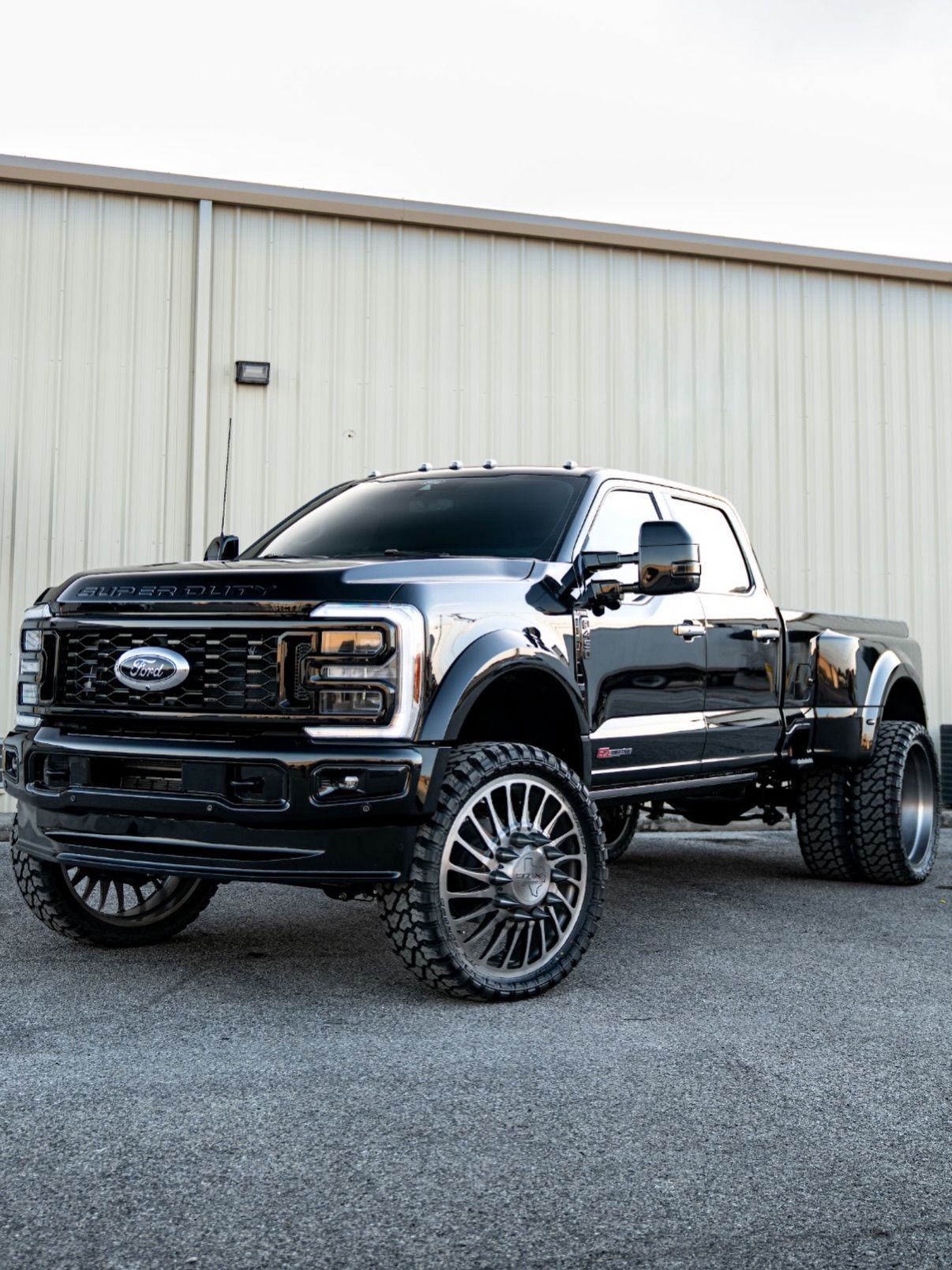 Black lifted Ford pickup truck parked on gravel in front of a beige metal building.