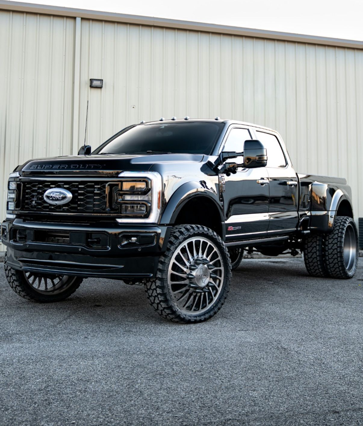 Black lifted Ford pickup truck with oversized wheels parked on gravel in front of a beige metal building
