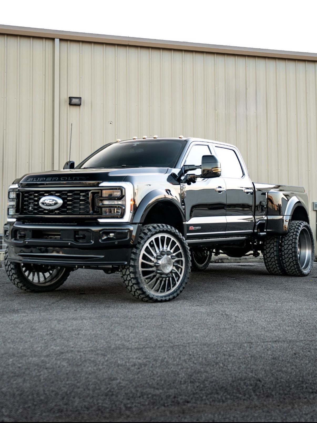 Black lifted Ford pickup truck parked on gravel in front of a beige industrial building