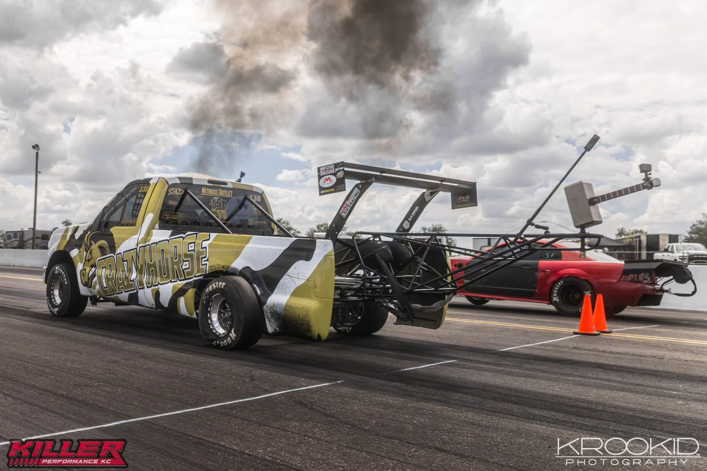 Two stripped race cars with smoke at a drag strip, one yellow and black, one red, with orange cones nearby