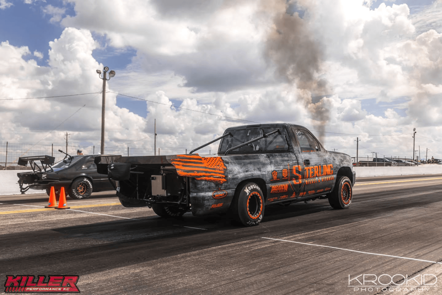 Two drag racing trucks at a track, one black with orange wheels launching from the старт line under cloudy skies