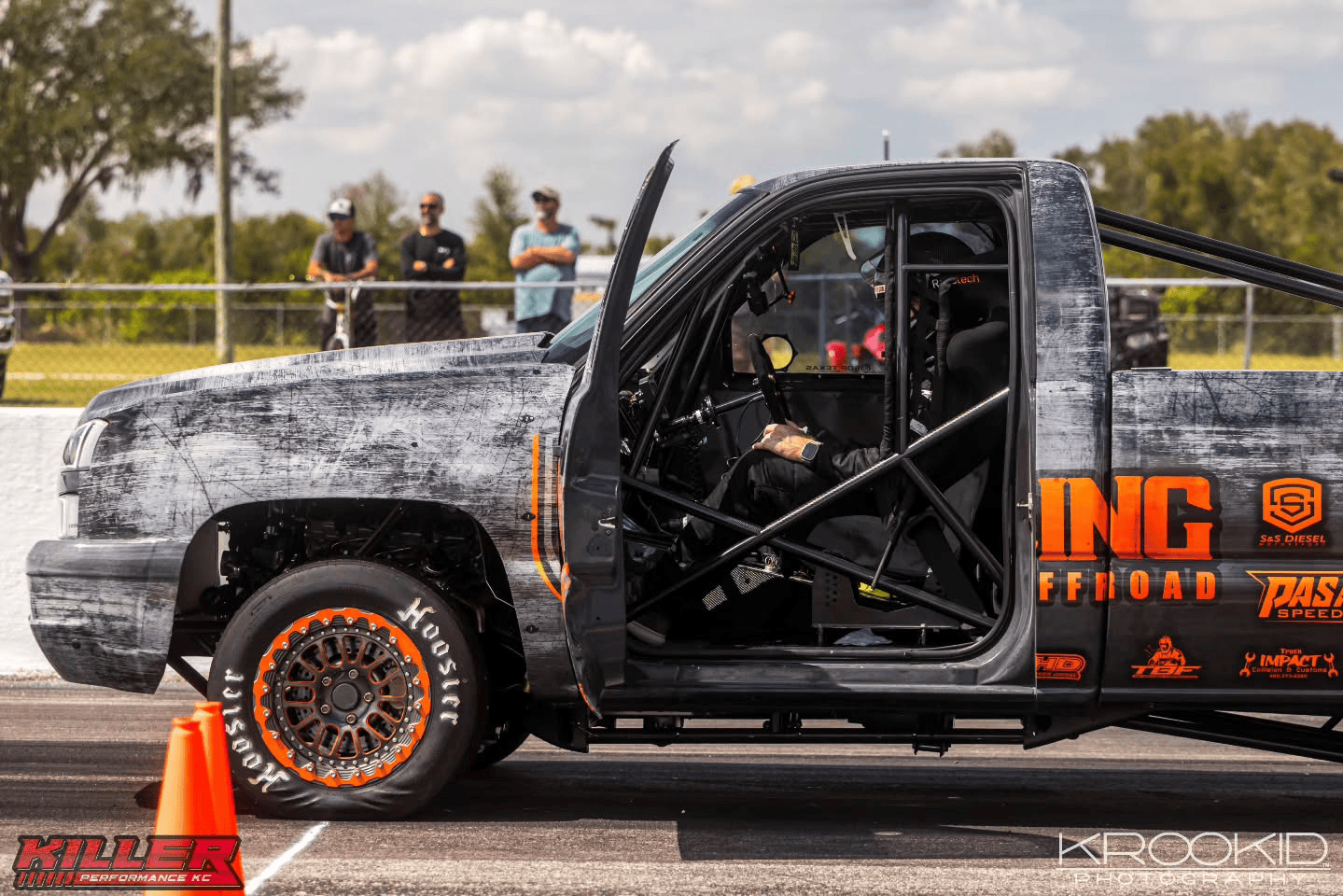 Black drag racing truck with roll cage and orange wheels staged on a track, spectators in the background