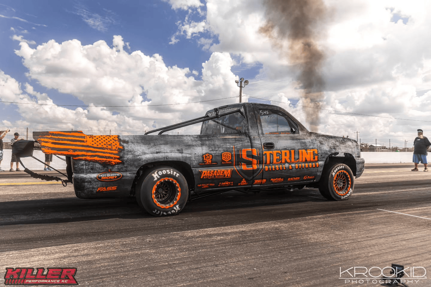 Black modified pickup truck with orange accents, burnout smoke, on a drag strip under cloudy sky