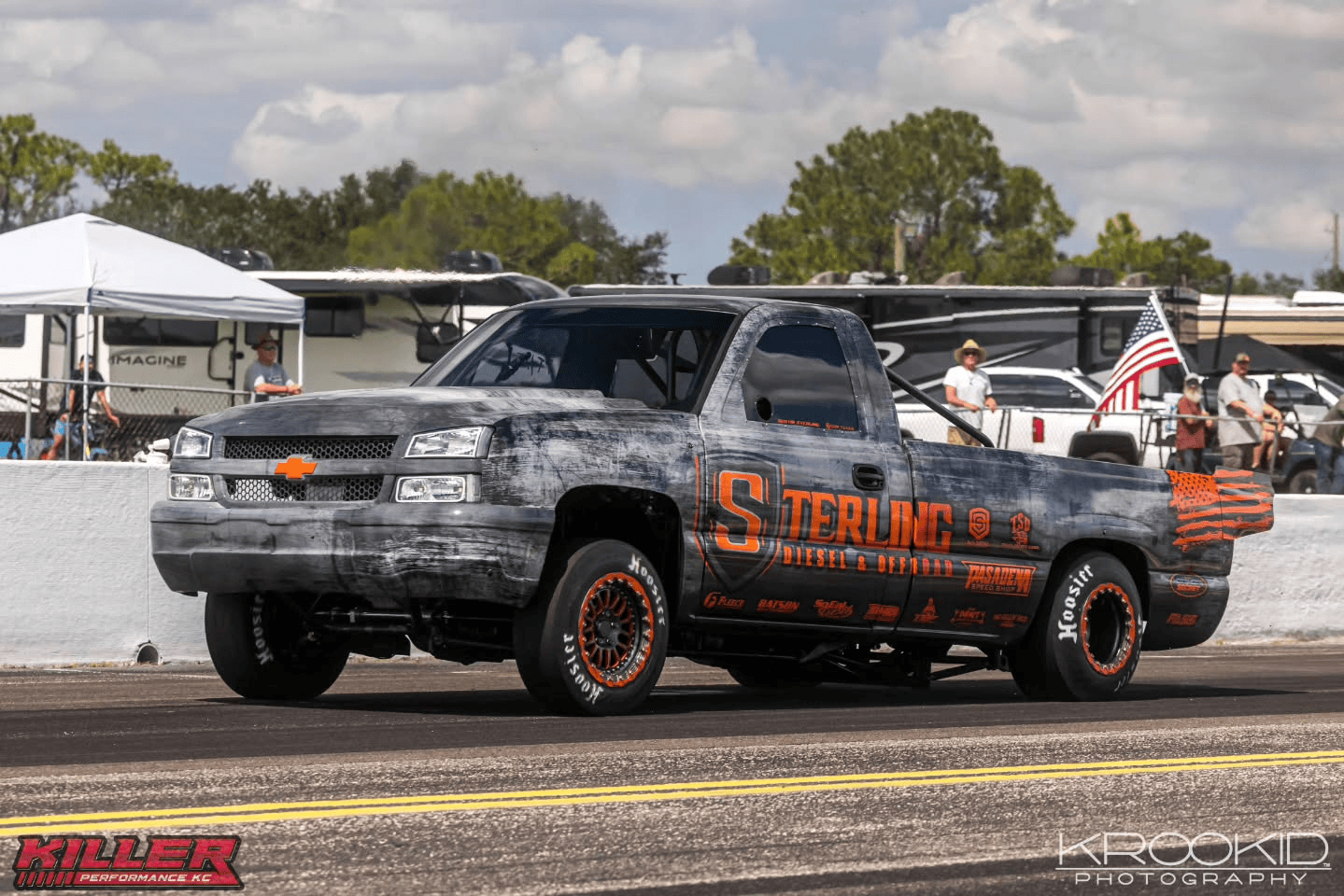 Camouflaged black-and-gray drag racing pickup truck on a track, with orange sponsor lettering and orange wheels