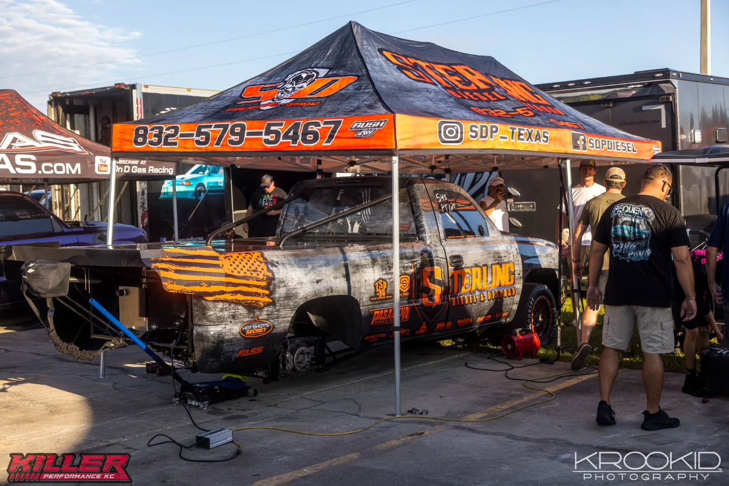 Orange-and-black motorsports tent with a customized car, equipment, and two people standing outside.