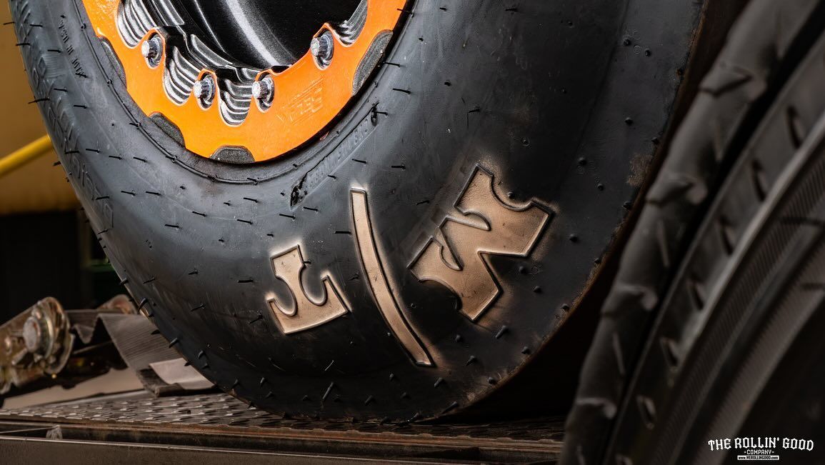 Close-up of a large off-road tire with orange rim and metal lettering on a dirt surface