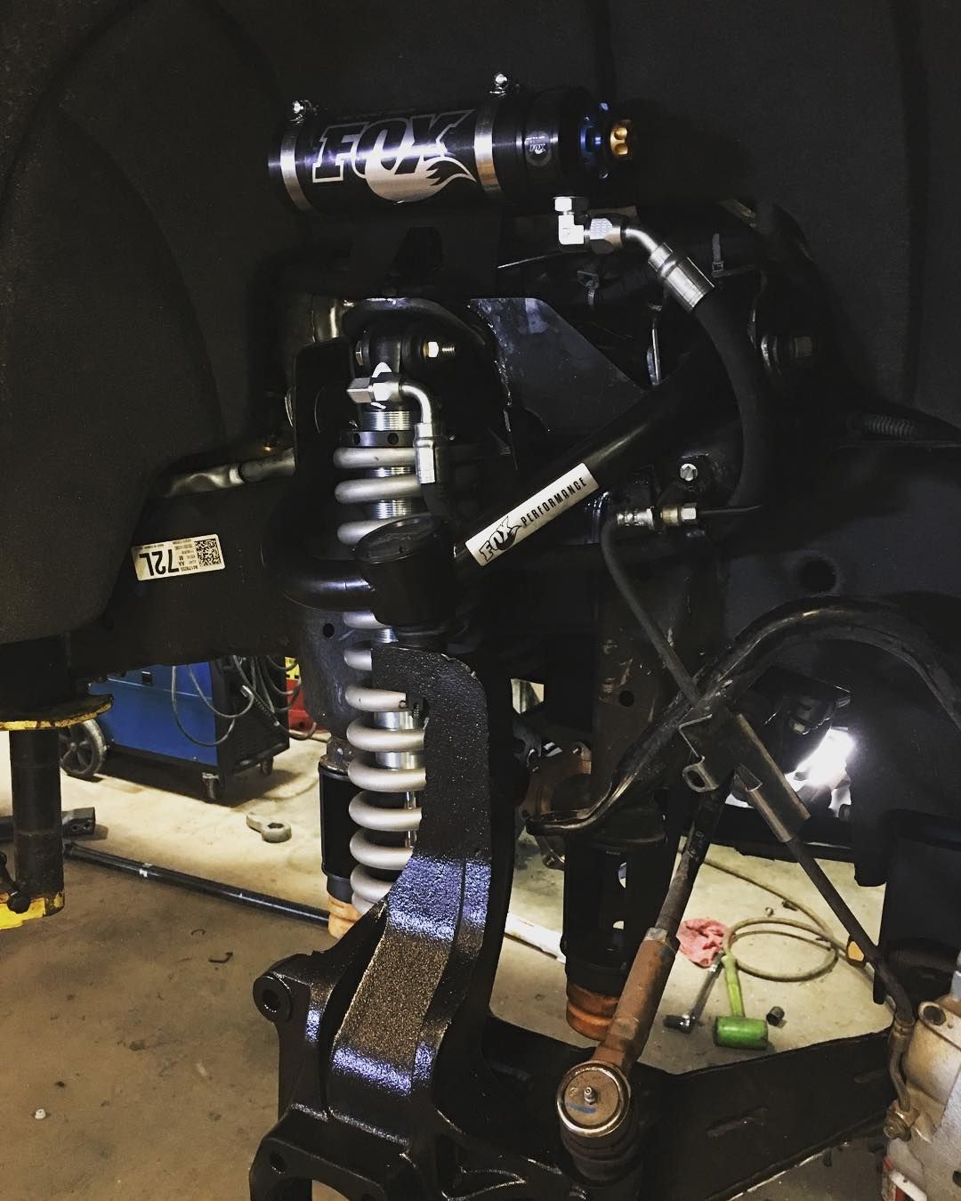 Black motorcycle suspension and frame parts in a dark garage, with a coil-over shock and exposed engine components.