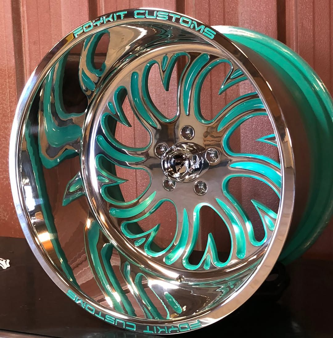 Turquoise custom chrome wheel rim with floral cutout design against a wood-paneled background