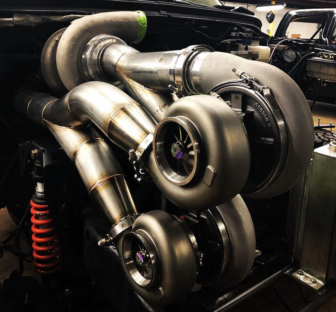 Close-up of a twin-turbo engine setup with polished metal piping in a dark workshop.