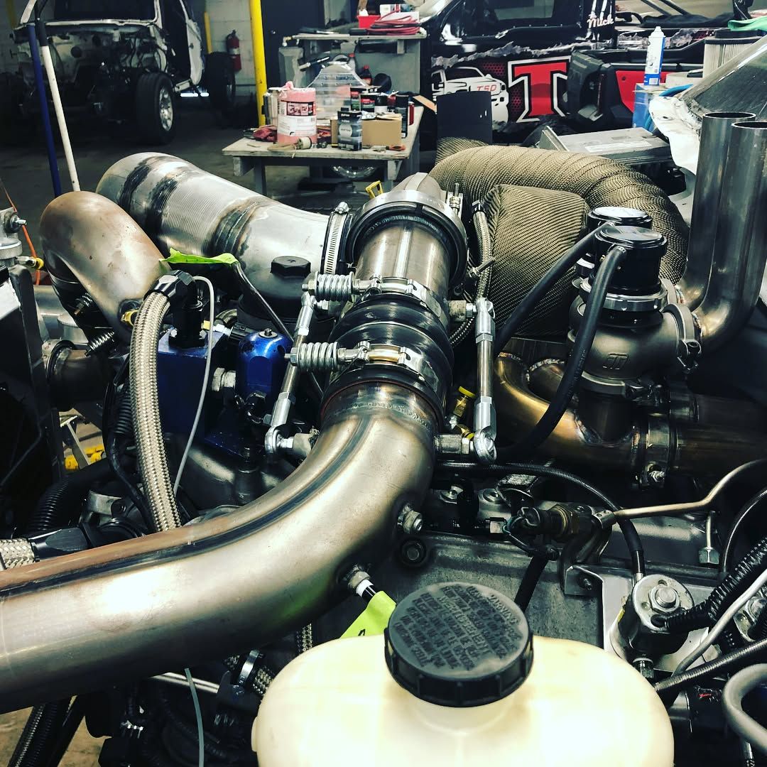Detailed car engine bay with shiny turbo piping, hoses, and mechanical components in a workshop.