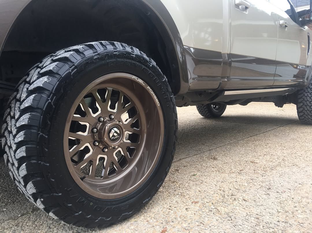 Close-up of a truck’s front wheel with a bronze rim and chunky off-road tire on a gravel surface