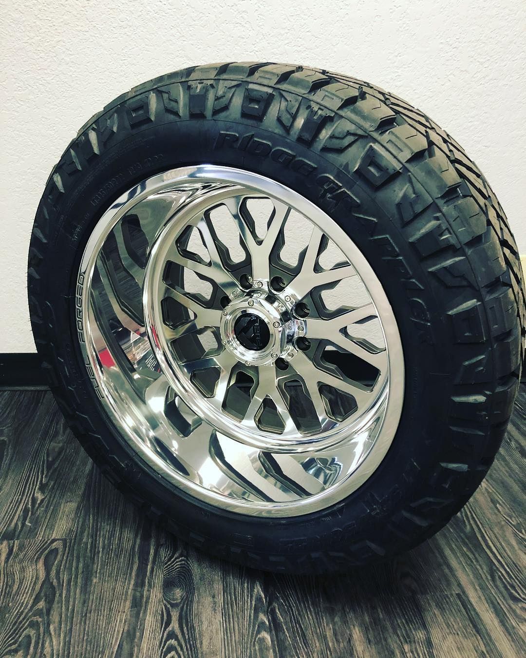 Chrome off-road wheel with black rugged tire, leaning against a wall on a wood floor.