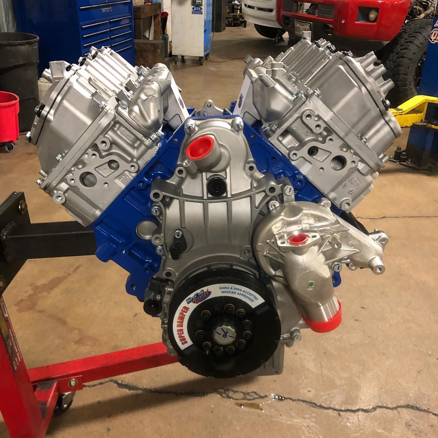 Blue and silver V8 engine mounted on a stand in a workshop, with red accents and exposed pulleys.