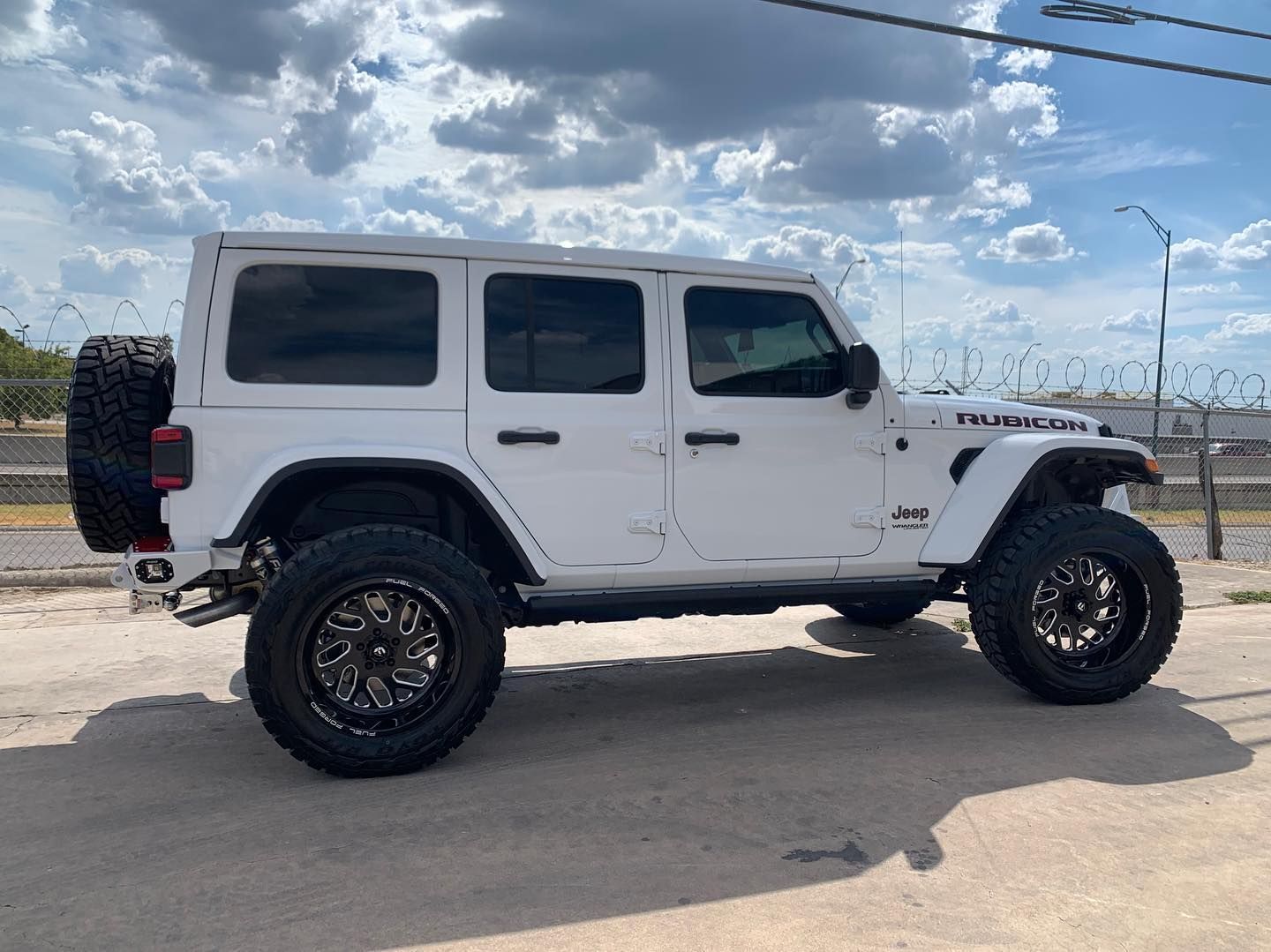 White Jeep Wrangler on a road under a cloudy sky, side view with lifted suspension and black wheels