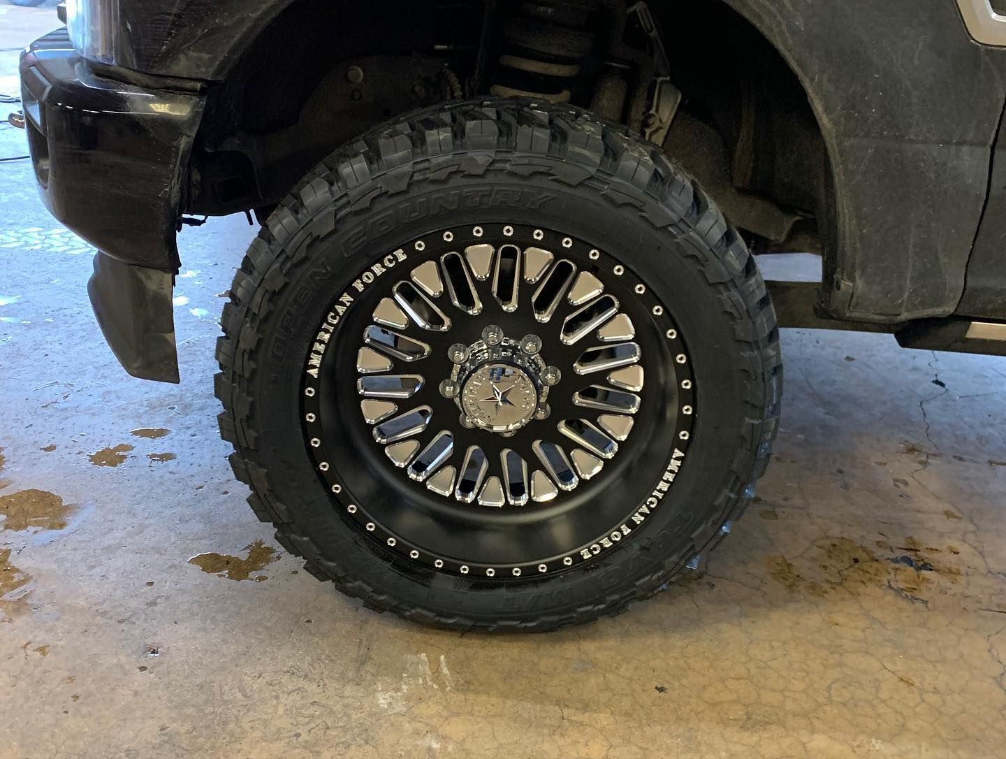 Black off-road truck tire and wheel on a wet garage floor, with a muddy fender above it