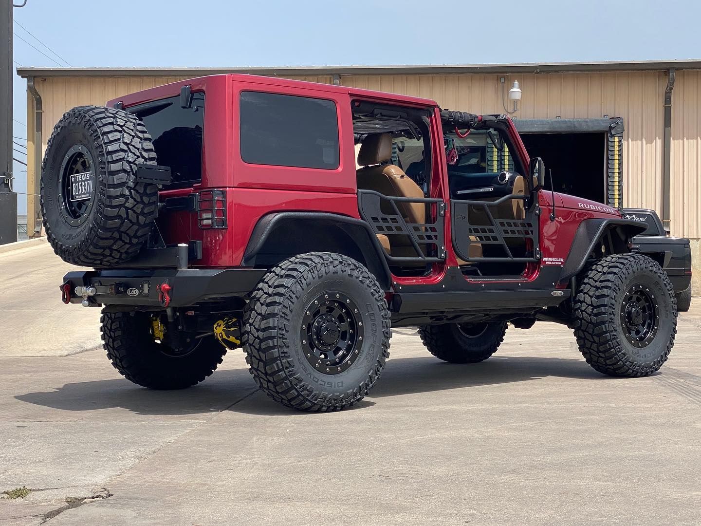 Red lifted Jeep Wrangler with open doors, large off-road tires, and rear spare tire parked on pavement.