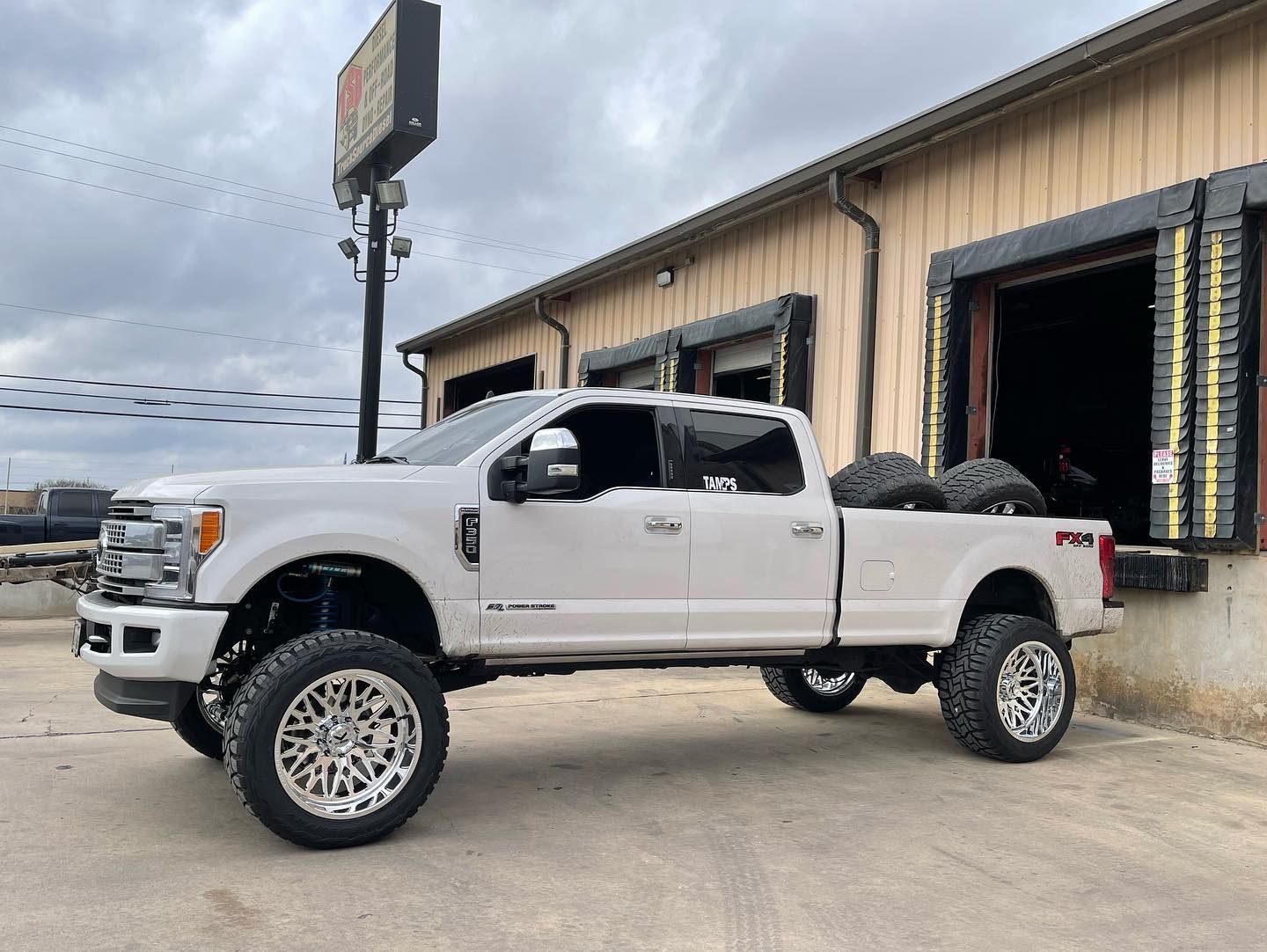 White lifted pickup truck parked beside a tan industrial building with open garage doors.