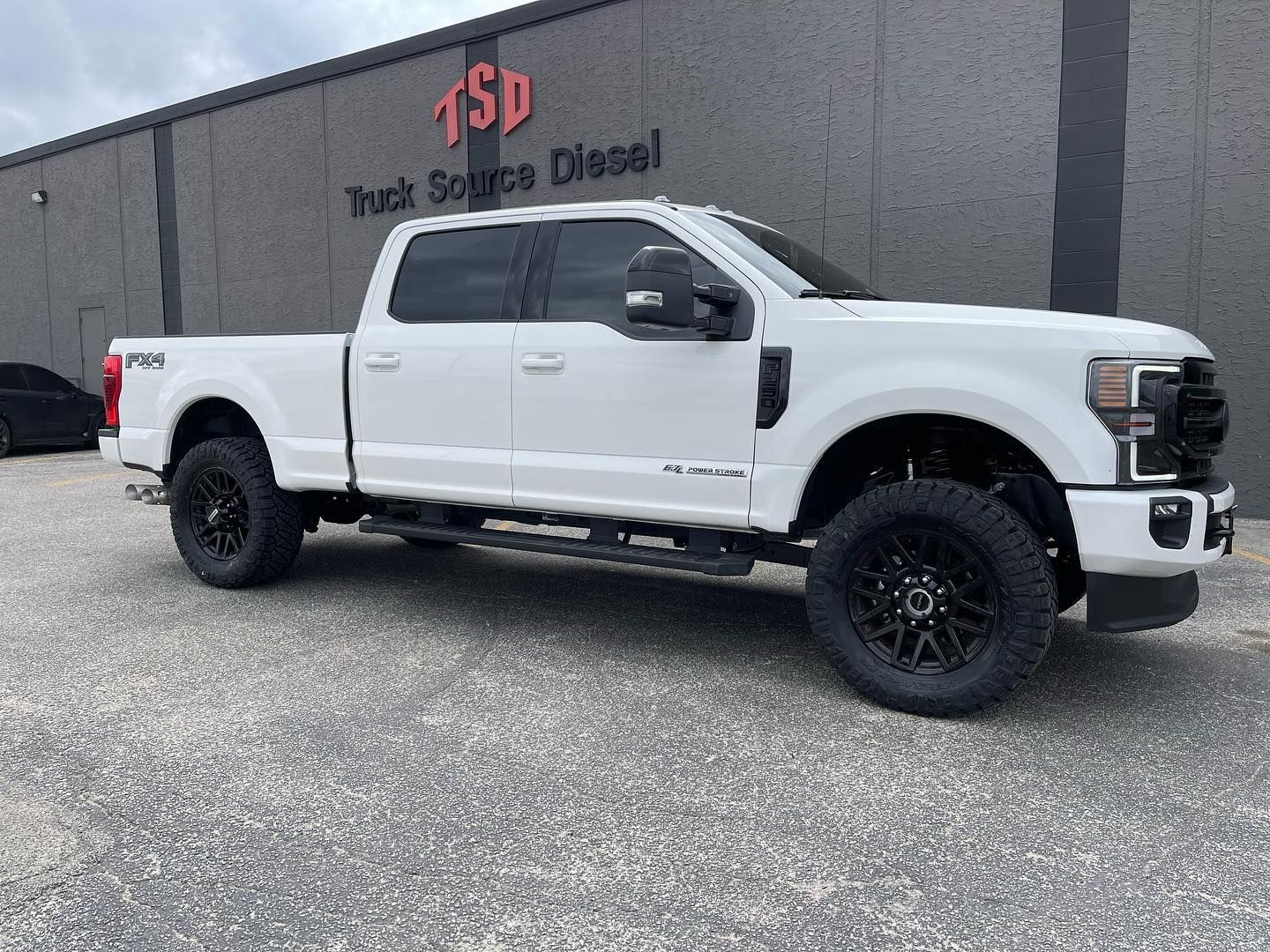 White lifted crew cab pickup parked outside TSD Truck Stuffing Diesel shop with black wheels and side steps