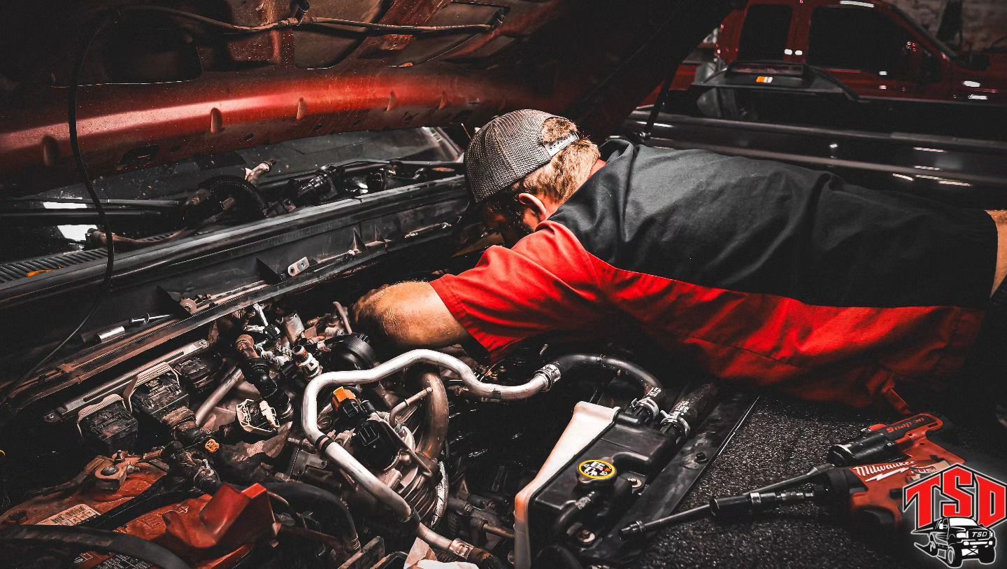Mechanic in red shirt working under a car hood in a dim garage with tools and engine parts visible