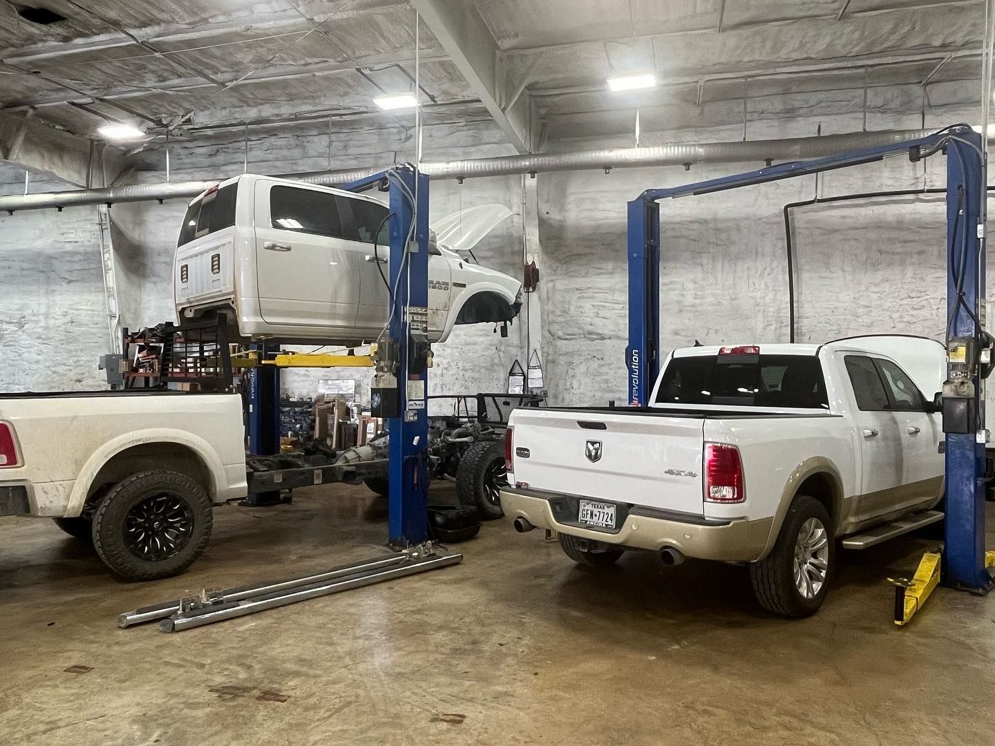 Two white pickup trucks in an auto shop with a raised truck on lifts and blue service posts