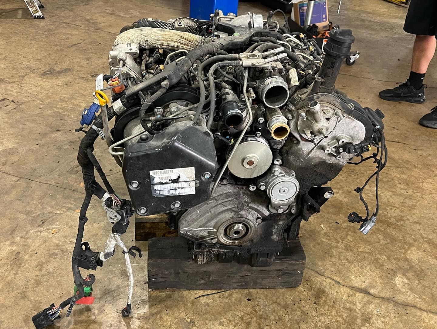 Detached car engine on a workshop floor, with tangled hoses and wires, near a person's legs.
