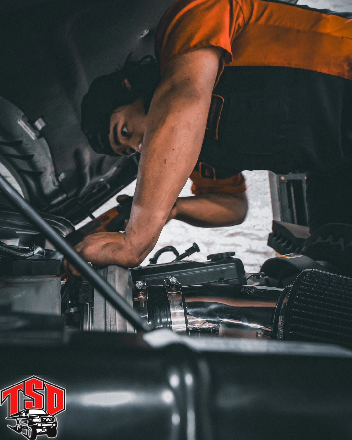 Mechanic in orange shirt working on car engine in a garage, leaning over with tools nearby