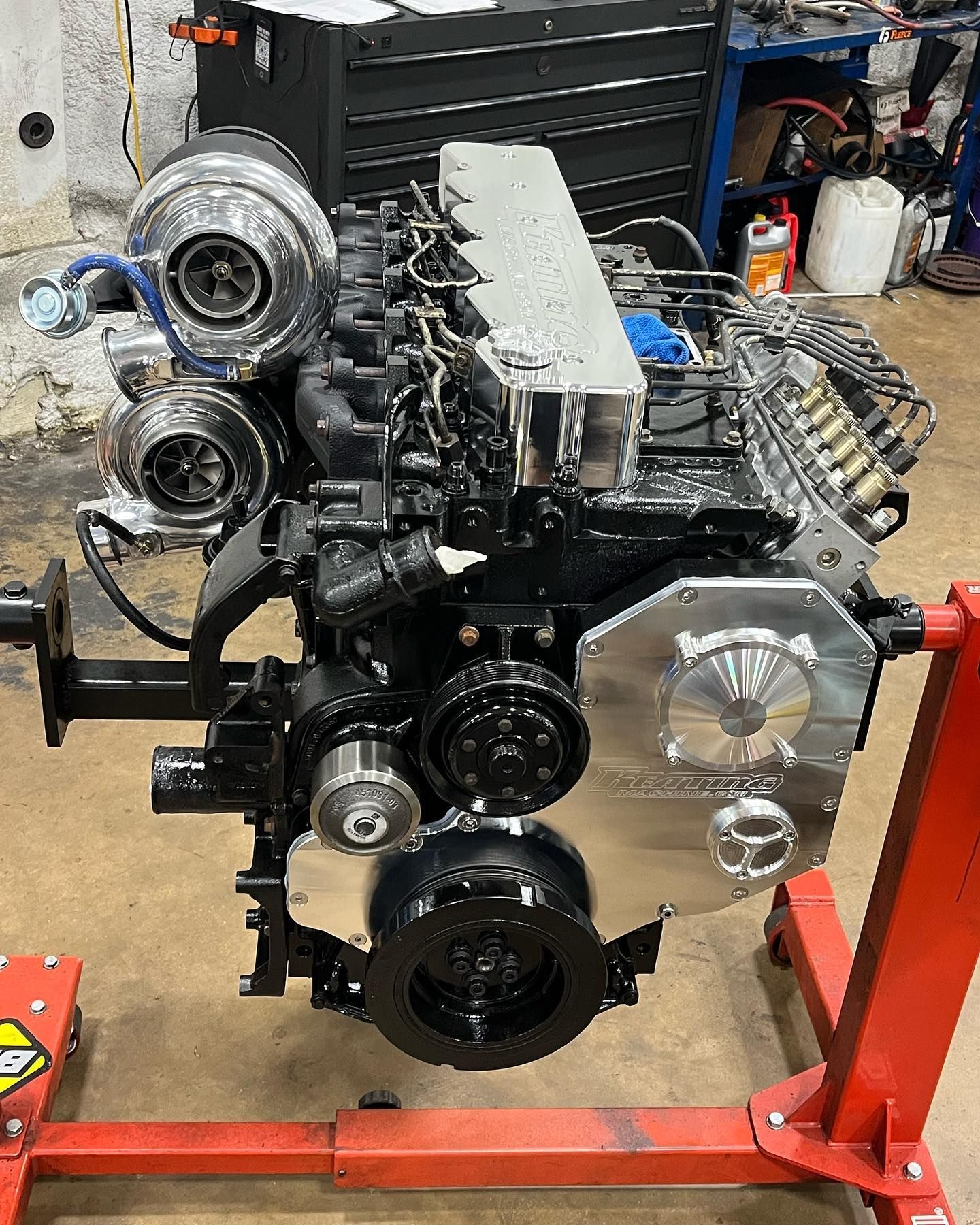 Large car engine mounted on a red stand in a workshop, with pulleys, belts, and exposed components visible.