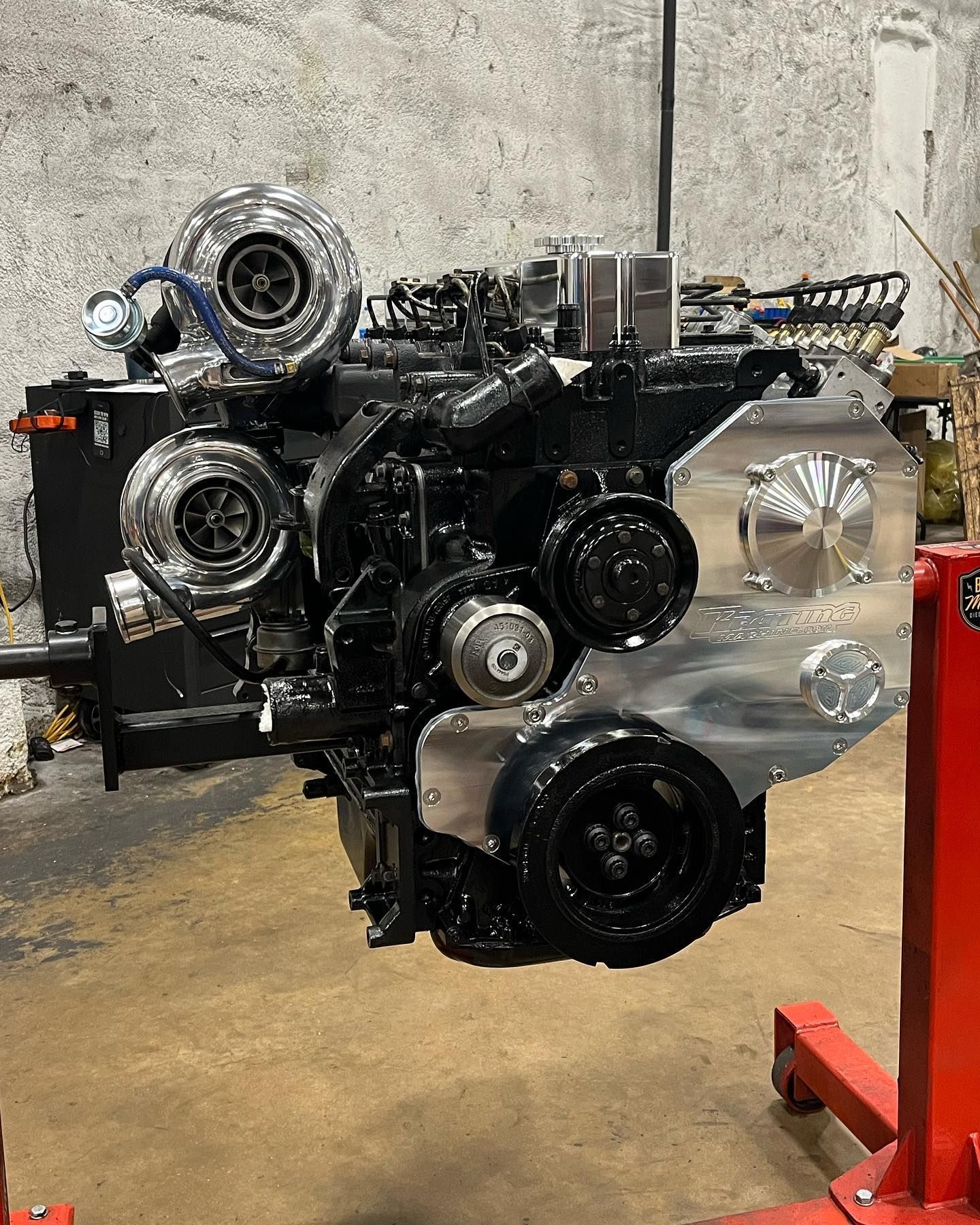 Black engine on a red stand in a workshop, with chrome pulleys and turbochargers visible.