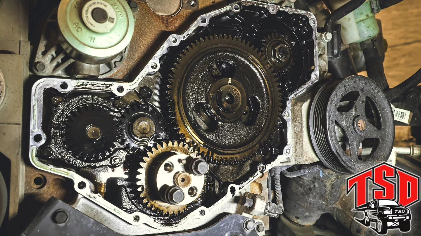 Open engine timing cover showing gears, chains, and oil-soaked metal parts.