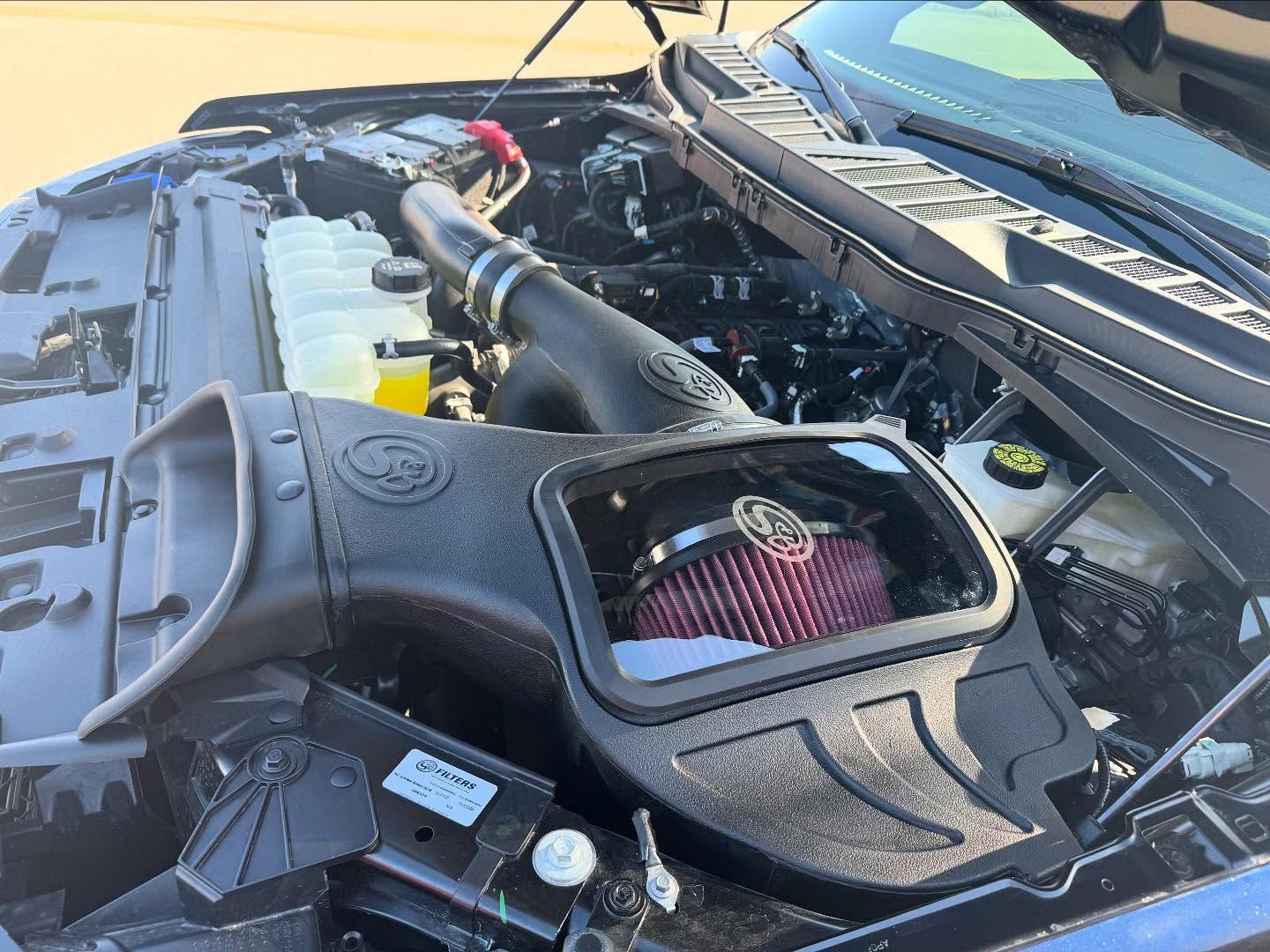 Open car engine bay with aftermarket air intake and yellow coolant reservoir at a sunny outdoor location