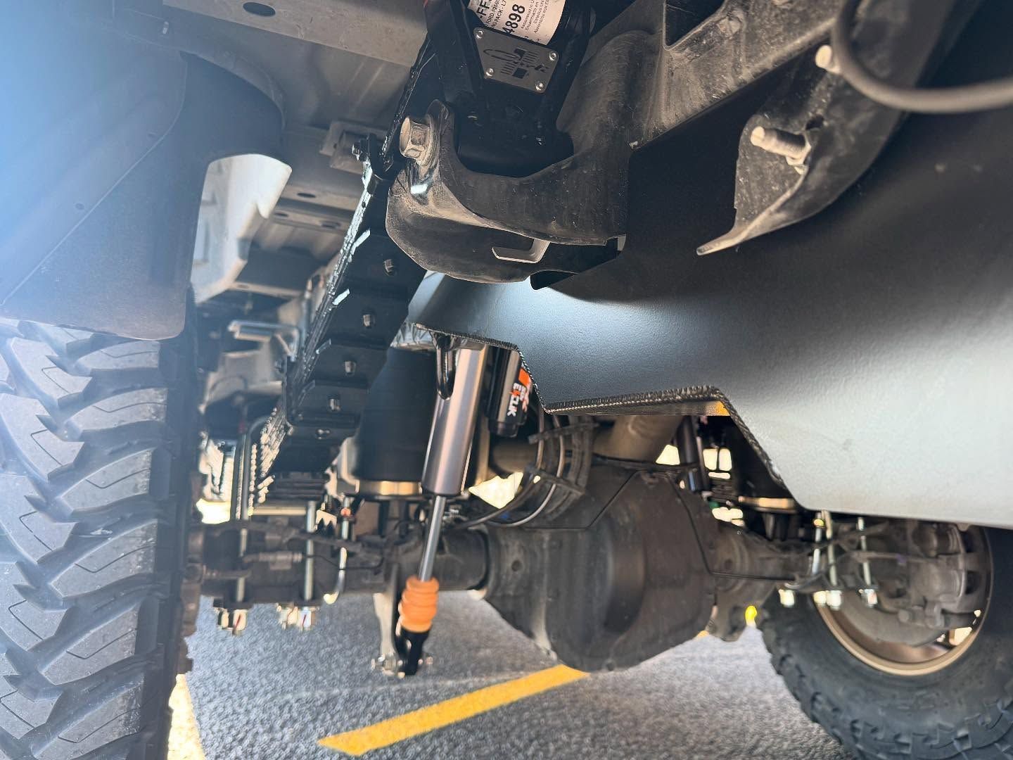 Undercarriage of a lifted off-road vehicle showing axle, suspension, and large tires on a road.
